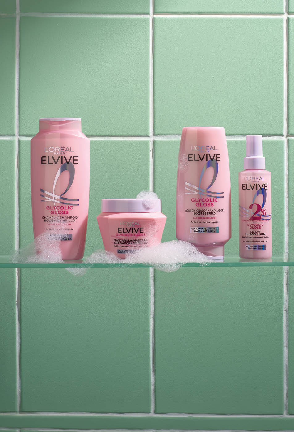 Cuatro productos de cuidado capilar L'Or al Elvive Glycolic Gloss en envases rosas sobre una repisa con burbujas y fondo de azulejos verdes. Descripci n generada con IA