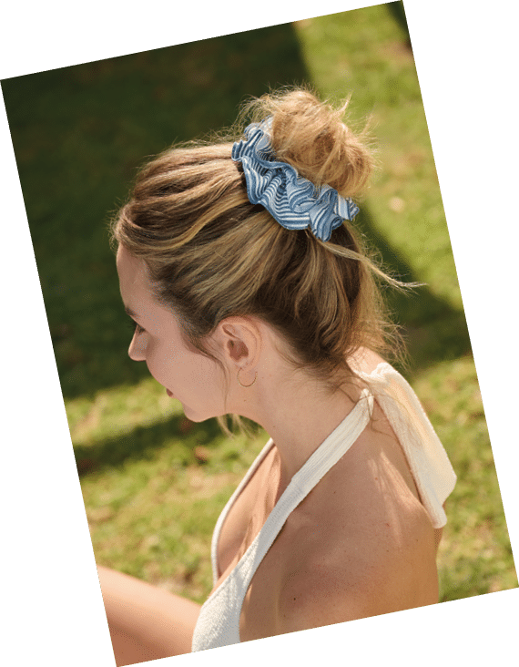 Persona con cabello recogido en un mo o sujetado con una scrunchie azul y blanca, vistiendo una prenda blanca sin mangas, sentada al aire libre sobre c sped. Descripci n generada con IA